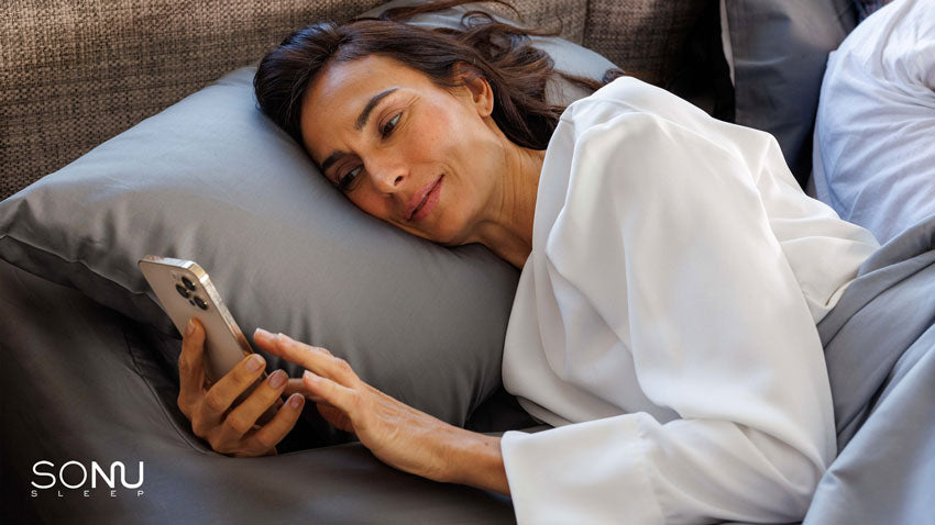 A photo of a female looking at her smart phone while resting on her side in a Sonu mattress with slate grey sheets.